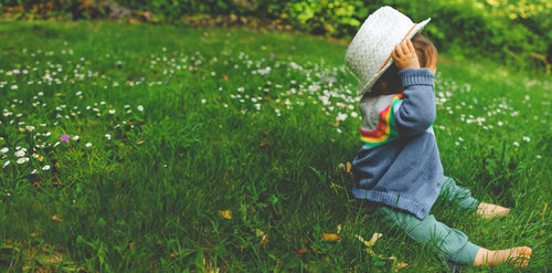 Little girl with a fun hat on sitting in the grass with pants and a sweater. Discover sustainable, affordable, gently-loved children's clothing bundles at Bundled, supporting parents and the environment with curated, high-quality options for newborns to toddlers at affordable and cheap price points.