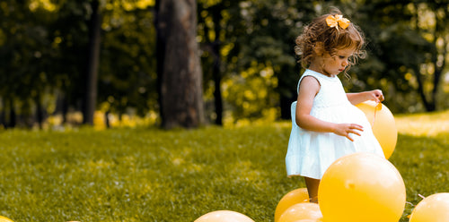 Little girl in a white dress playing outside with yellow balloons. Discover sustainable, affordable, gently-loved children's clothing bundles at Bundled, supporting parents and the environment with curated, high-quality options for newborns to toddlers at affordable and cheap price points.