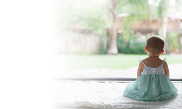 Little girl sitting in a white and green dress, looking outside at the greenery. Discover sustainable, affordable, gently-loved children's clothing bundles at Bundled, supporting parents and the environment with curated, high-quality options for newborns to toddlers at affordable and cheap price points.