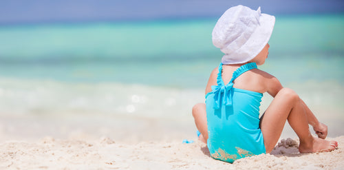 Little girl in a blue bathing suit sitting on the beach looking at the water while playing in the sand. Discover sustainable, affordable, gently-loved children's clothing bundles at Bundled, supporting parents and the environment with curated, high-quality options for newborns to toddlers at affordable and cheap price points.